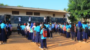 Alunos uniformizados reúnem-se no pátio de uma escola moçambicana para o início das aulas, enquanto professores orientam a formação, simbolizando disciplina e compromisso com a educação.